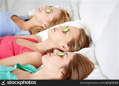 people, beauty, natural cosmetics and pajama party concept - happy friends or teenage girls with cucumber mask lying in bed at home