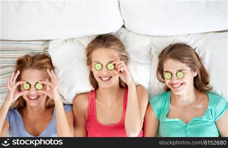people, beauty, natural cosmetics and pajama party concept - happy friends or teenage girls with cucumber mask having fun and lying in bed at home