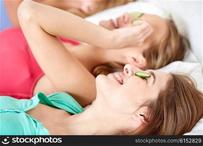 people, beauty, natural cosmetics and pajama party concept - happy friends or teenage girls with cucumber mask having fun and lying in bed at home