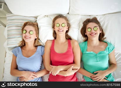 people, beauty, natural cosmetics and pajama party concept - happy friends or teenage girls with cucumber mask having fun and lying in bed at home