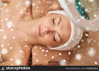 people, beauty, skin care, winter and relaxation concept - close up of beautiful young woman face with closed eyes under magnifying lamp in spa salon with snow effect