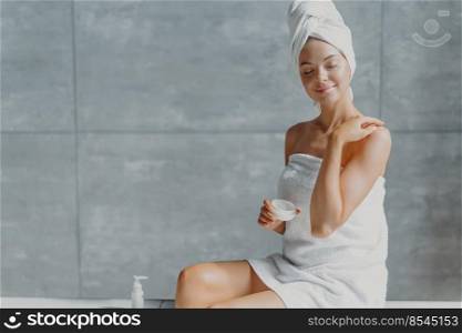 People, beauty, spa and cosmetology concept. Relaxed young European female puts body cream, touches shoulder gently, wrapped in white soft towel, closes eyes with pleasure, poses against grey wall