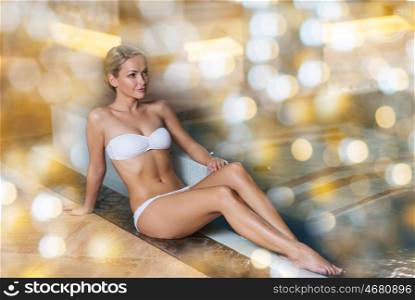 people, beauty, spa and relaxation concept - beautiful young woman in bikini swimsuit sitting on swimming pool edge over holidays lights background