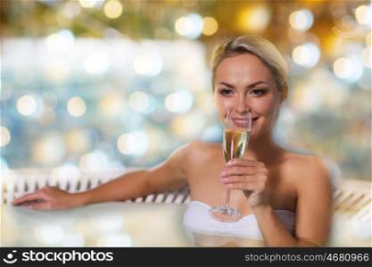people, beauty, spa and relaxation concept - beautiful young woman wearing bikini swimsuit sitting with glass of champagne in jacuzzi at poolside over holidays lights background
