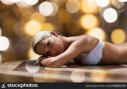 people, beauty, spa, bodycare and relaxation concept - beautiful young woman lying on hammam table in turkish bath over holidays lights background