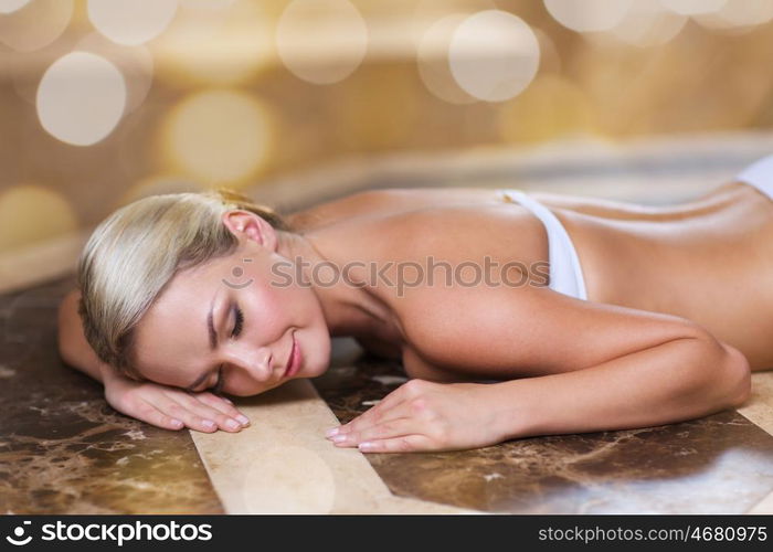 people, beauty, spa, bodycare and relaxation concept - beautiful young woman lying on hammam table in turkish bath over holidays lights background