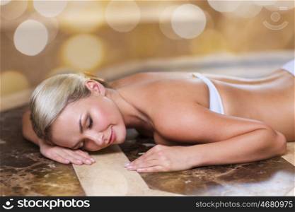 people, beauty, spa, bodycare and relaxation concept - beautiful young woman lying on hammam table in turkish bath over holidays lights background