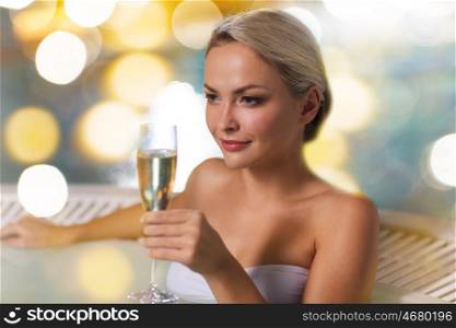 people, beauty, spa, healthy lifestyle and relaxation concept - beautiful young woman wearing bikini swimsuit sitting with glass of champagne in jacuzzi at poolside over holidays lights background
