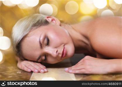 people, beauty, spa, healthy lifestyle and relaxation concept - beautiful young woman lying on hammam table in turkish bath over holidays lights background