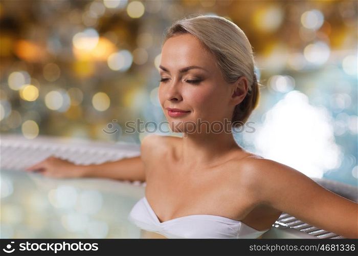 people, beauty, spa, healthy lifestyle and relaxation concept - beautiful young woman wearing bikini swimsuit sitting in jacuzzi at poolside over holidays lights background