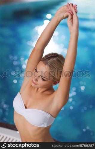 people, beauty, spa, healthy lifestyle and relaxation concept - beautiful young woman wearing bikini swimsuit sitting with raised hands in jacuzzi at poolside