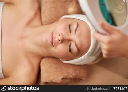 people, beauty, spa, healthy lifestyle and relaxation concept - close up of beautiful young woman lying with closed eyes and cosmetologist looking through magnifying lamp in spa
