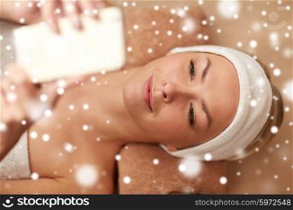 people, beauty, spa, technology and winter concept - close up of beautiful smiling young woman lying with smartphone in spa salon with snow effect