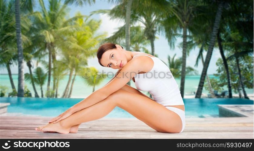 people, beauty, spa, travel and resort concept - beautiful woman in cotton underwear touching her legs over swimming pool on beach with palms background