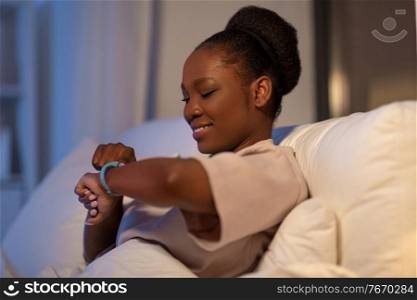 people, bedtime and rest concept - african american woman looking at health tracker in bed at home at night. woman with health tracker in bed at night