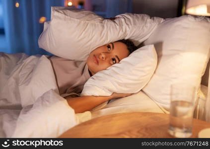 people, bedtime and rest concept - annoyed african american woman lying in bed at night and covering ears with pillow. angry african woman lying in bed and closing ears