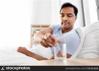 people, bedtime and rest concept - happy indian man lying in bed at home and taking glass of water from table. happy indian man drinking water lying in bed