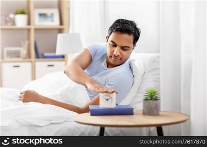 people, bedtime and rest concept - happy indian man with alarm clock lying in bed at home. happy indian man with alarm clock lying in bed