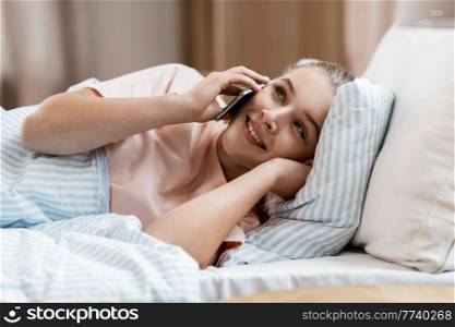 people, bedtime and rest concept - happy smiling girl calling on smartphone lying in bed at home. happy girl calling on smartphone in bed at home