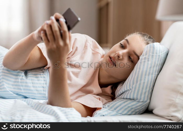 people, bedtime and rest concept - happy smiling girl with smartphone lying in bed at home. happy girl with smartphone in bed at home