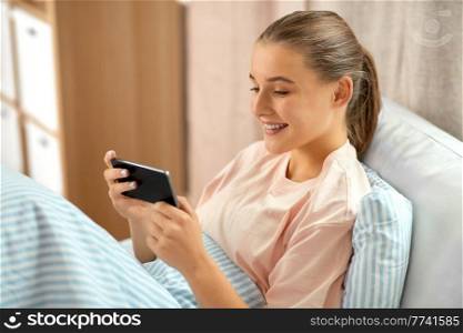 people, bedtime and rest concept - happy smiling girl with smartphone lying in bed at home. happy girl with smartphone in bed at home