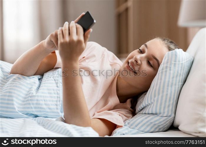 people, bedtime and rest concept - happy smiling girl with smartphone lying in bed at home. happy girl with smartphone in bed at home