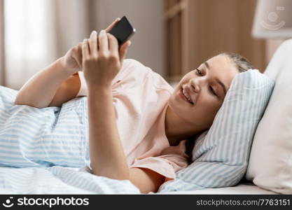 people, bedtime and rest concept - happy smiling girl with smartphone lying in bed at home. happy girl with smartphone in bed at home