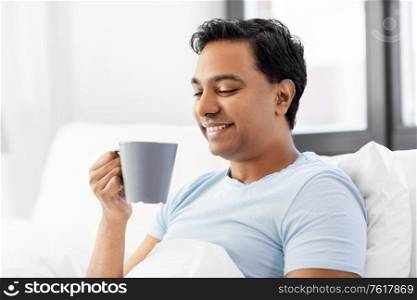 people, bedtime and rest concept - happy smiling indian man drinking coffee in bed at home. happy indian man drinking coffee in bed at home