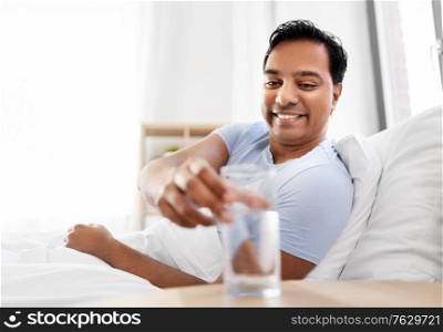 people, bedtime and rest concept - happy smiling indian man lying in bed at home and taking glass of water from table. happy indian man drinking water lying in bed