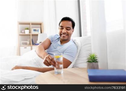 people, bedtime and rest concept - happy smiling indian man lying in bed at home and taking glass of water with lemon from table. happy indian man drinking water lying in bed