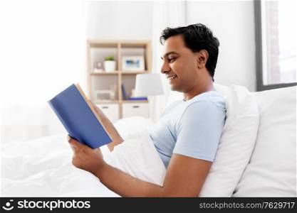 people, bedtime and rest concept - happy smiling indian man reading book in bed at home. happy indian man reading book in bed at home