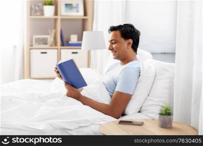 people, bedtime and rest concept - happy smiling indian man reading book in bed at home. happy indian man reading book in bed at home