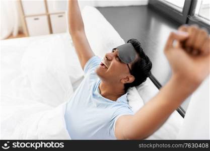 people, bedtime and rest concept - happy smiling indian man with eye sleeping mask stretching in bed at home. happy smiling indian man lying in bed at home