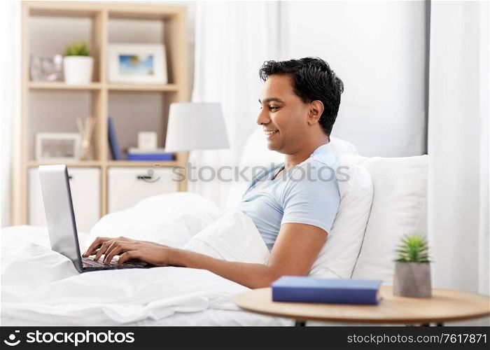 people, bedtime and rest concept - happy smiling indian man with laptop computer lying in bed at home. happy indian man with laptop in bed at home