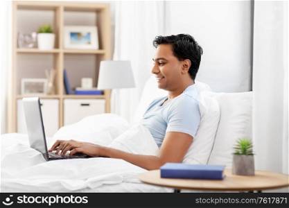 people, bedtime and rest concept - happy smiling indian man with laptop computer lying in bed at home. happy indian man with laptop in bed at home