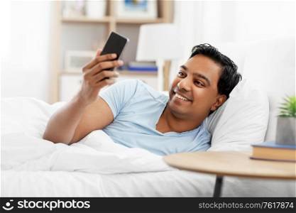 people, bedtime and rest concept - happy smiling indian man with smartphone lying in bed at home. happy indian man with smartphone in bed at home