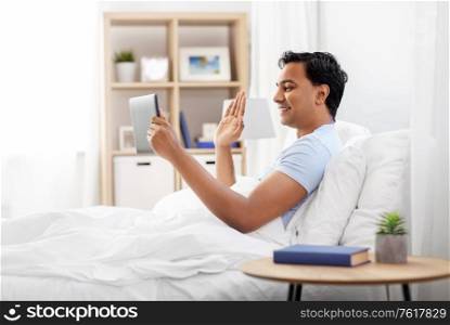 people, bedtime and rest concept - happy smiling indian man with tablet pc computer lying in bed and having video call at home. happy man with tablet pc in bed having video call