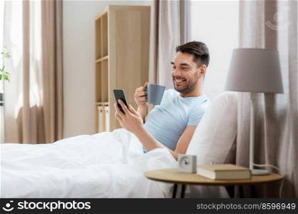 people, bedtime and rest concept - happy smiling man with smartphone drinking coffee in bed at home. happy man with smartphone in bed at home
