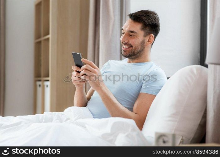 people, bedtime and rest concept - happy smiling man with smartphone lying in bed at home. happy man with smartphone in bed at home