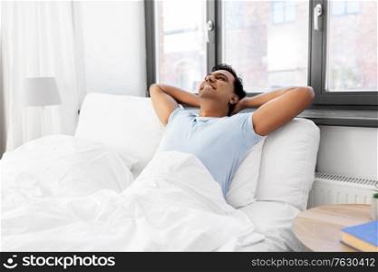 people, bedtime and rest concept - happy smiling relaxed indian man stretching in bed at home. happy smiling indian man stretching in bed at home