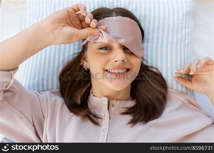 people, bedtime and rest concept - happy smiling teenage girl in eye sleeping mask lying in bed at home in morning. happy girl in eye sleeping mask lying in bed