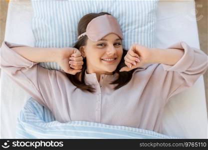 people, bedtime and rest concept - happy smiling teenage girl in eye sleeping mask stretching in bed at home in morning. girl in eye sleeping mask stretching in bed