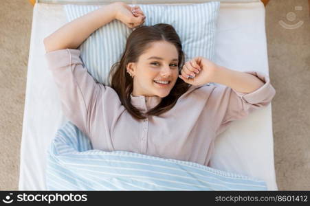 people, bedtime and rest concept - happy smiling teenage girl stretching in bed at home in morning. happy smiling girl stretching in bed