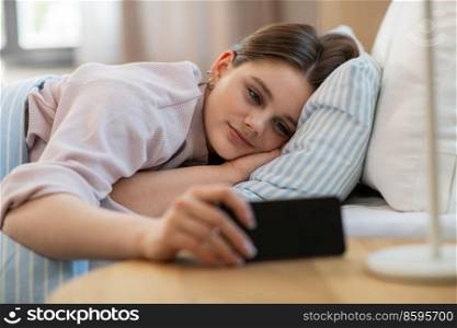 people, bedtime and rest concept - happy smiling teenage girl with smartphone lying in bed at home. happy girl with smartphone in bed at home