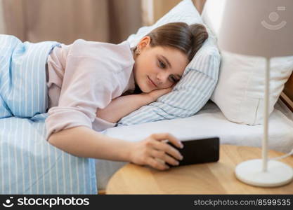 people, bedtime and rest concept - happy smiling teenage girl with smartphone lying in bed at home. happy girl with smartphone in bed at home