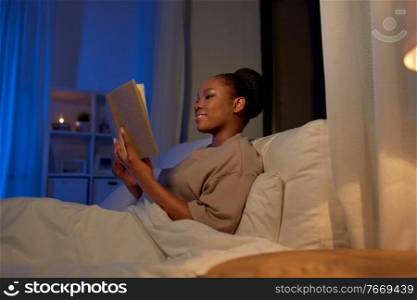 people, bedtime and rest concept - happy smiling young african american woman reading book in bed at home at night. young woman reading book in bed at home