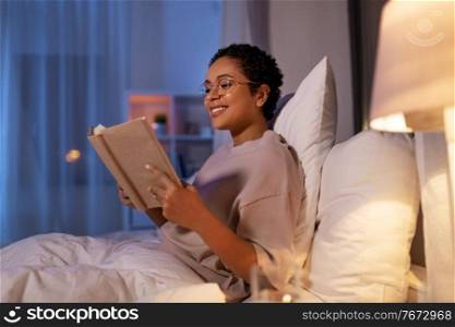 people, bedtime and rest concept - happy smiling young african american woman in glasses reading book in bed at home at night. smiling young woman reading book in bed at home