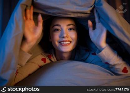 people, bedtime and rest concept - happy teenage girl lying under blanket in bed at home at night. happy teenage girl lying under blanket in bed
