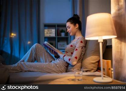 people, bedtime and rest concept - happy teenage girl reading book in bed at home at night. happy teenage girl reading book in bed at home