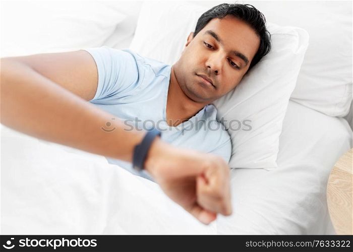 people, bedtime and rest concept - indian man looking at health tracker in bed at home. indian man with health tracker in bed at home